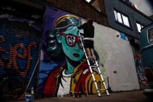 Street artist on a ladder painting a colourful portrait mural in Hackney Wick