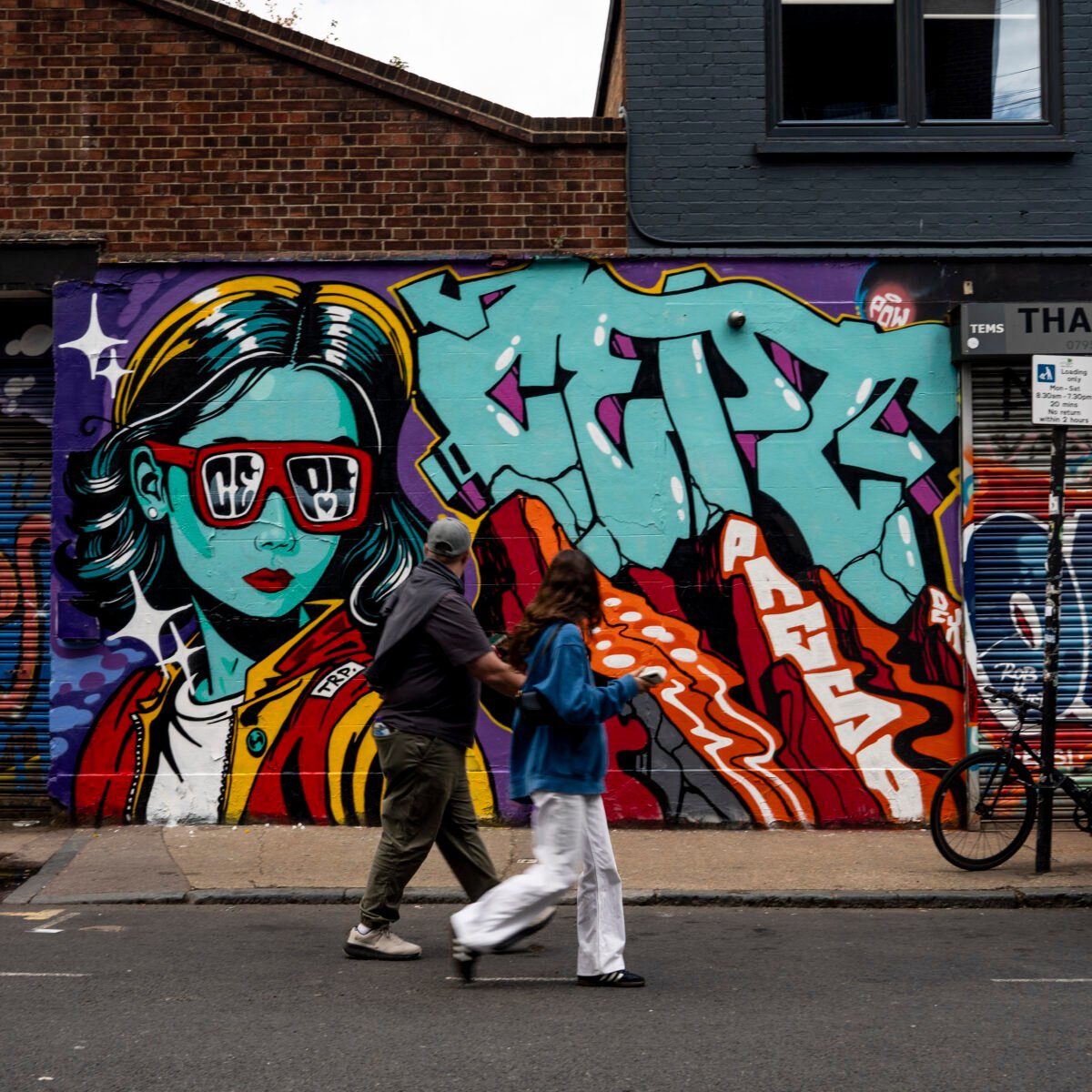 Two pedestrians walking past a colourful street-art mural on White Post Lane in Hackney Wick