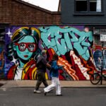 Two pedestrians walking past a colourful street-art mural on White Post Lane in Hackney Wick